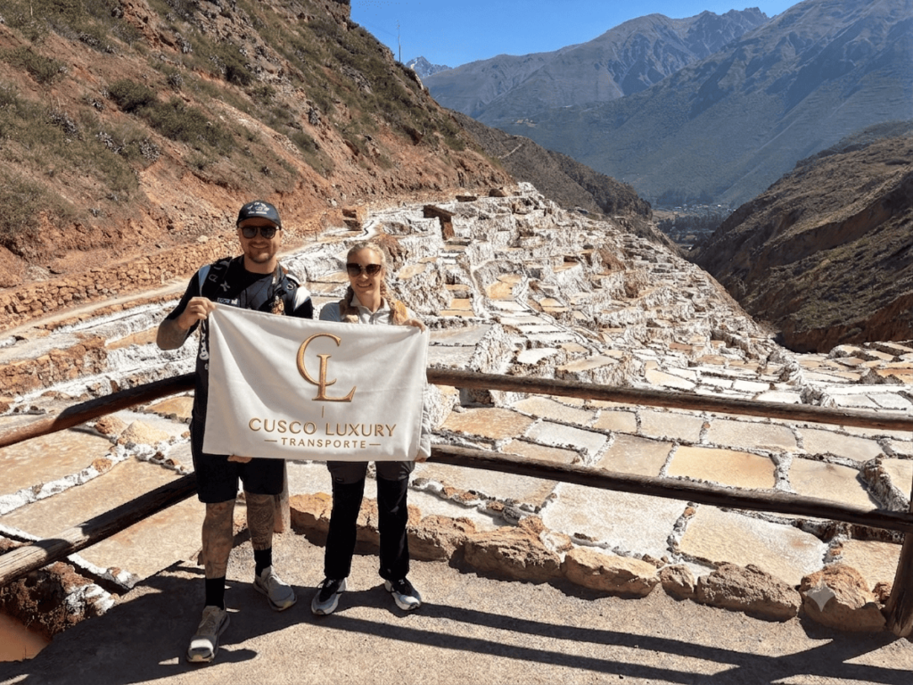 Two smiling tourists holding a Cusco Luxury Transporte flag at the Maras Salt Mines viewpoint during a Sacred Valley stopover transfer.